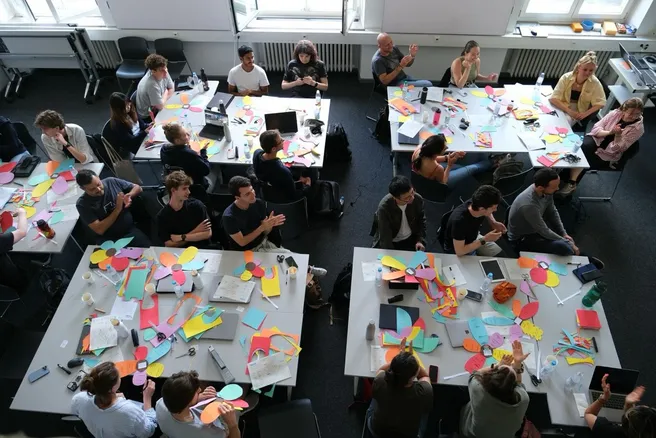 A view from above: Students sitting in a room on group tables and working on a workshop called "Flowers of Proximity"